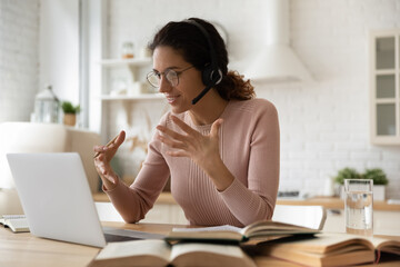 Young woman college student e-learn sit in kitchen wear headset look at laptop talk to tutor use videoconference, heap of textbooks on table. Digital online educational course, videocall event concept