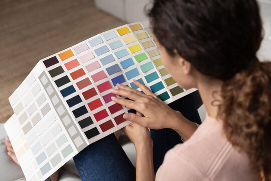 Rear close up view over female shoulder holds swatches looks at pantone samples, choose colors for coming repairs renovation works at home. Designer work indoor holds palette set make shades selection