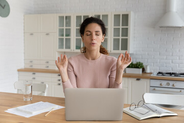 Young Hispanic businesswoman sit at table in kitchen near computer closed eyes makes breathing...