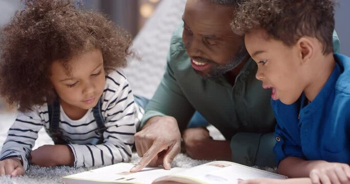 Close up of happy grandfather lying on the floor with two grandchildren, reading them book flipping through pages, looking at pictures.