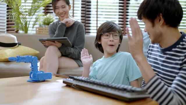 STEM Education for kids.A nerd in glasses is holding a robot. Asian boy in a green shirt and father is sitting at table in front of him whose robot arm the boy made.He is very satisfied with his work