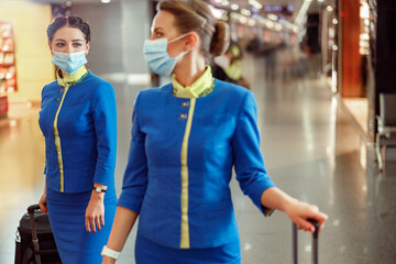 Women stewardesses wearing protective face masks and air hostess uniform while walking down airport terminal during pandemic