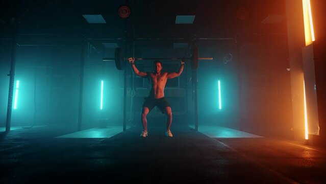 Male powerlifter in a blue-orange neon light is preparing for a workout in the gym. A strong man with a naked torso does lifting a barbell over his head, a jerk exercise from weightlifting.