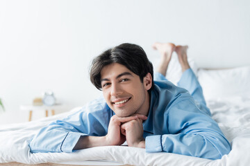 young brunette man in blue pajamas lying on bed and smiling at camera.