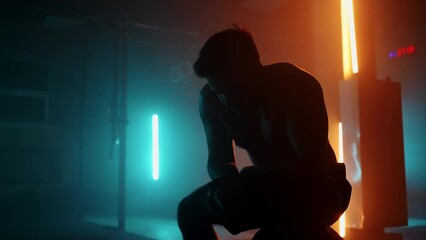 A tired athlete is resting in breathing heavily while sitting on a barbell in neon light after performing an exercise in slow motion. Recuperate and prepare for a new breakthrough