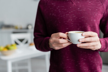 partial view of man holding cup of morning coffee in blurred kitchen.