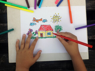 A child`s hands grab a red marker and draw.
Drawing made by a kid. Children`s illustration of a house. A garden with colorful flowers, grass and a tree. A sky with clouds, a plane and a smiling sun.