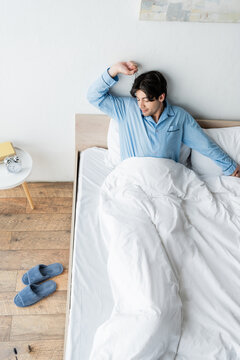 High Angle View Of Man In Blue Pajamas Stretching In Bed Near Vintage Alarm Clock On Bedside Table.