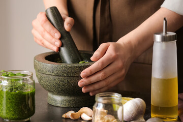 Woman making pesto sauce with mortar and pestle on table in kitchen - obrazy, fototapety, plakaty
