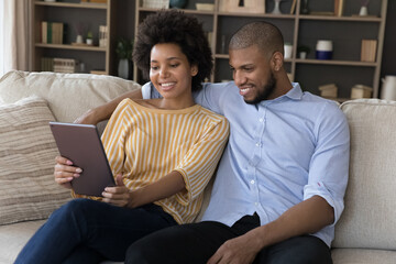 Beautiful African couple relaxing on sofa using digital tablet, choosing online goods, buying e-services at home, booking hotel planning vacation. Modern tech, distancing services, app usage concept