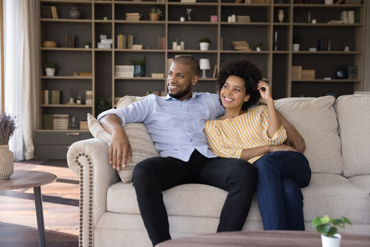 Beautiful African couple in love relax on couch in fashionable living room smile looking into distance, spouses enjoy weekend at own or rented house, daydreaming feels happy. Homeowners family concept