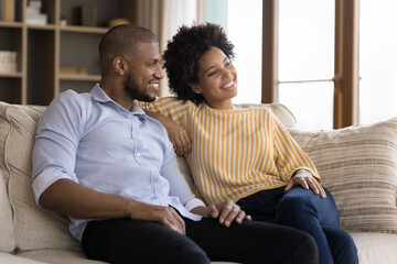 Dreamy African couple in love sit on sofa in living room, daydreaming looking into distance smile...