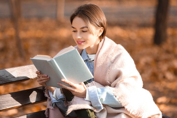 Obraz premium Young woman with plaid reading book on wooden bench in autumn park