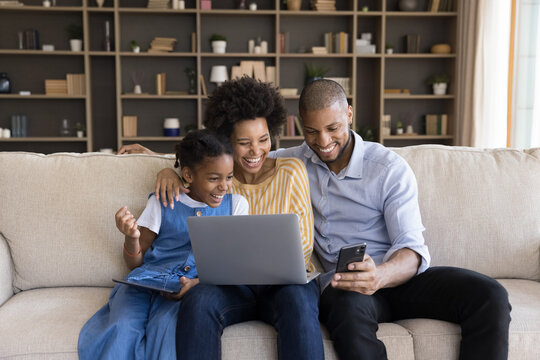 Young parents and little 7s daughter sit on sofa with diverse devices watching on cellphone funny videos laughing looking overjoyed. African family enjoy modern tech, online humorous content concept