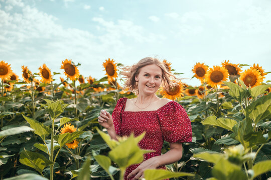 Woman in red summer style dress with open hands over cloudy blue sky. Beautiful caucasian girl with long blond hair in a blooming sunflower field.