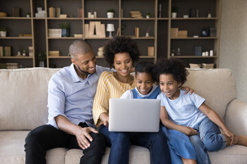Young African couple with kids sit on sofa watch family movie online on laptop, use digital streaming services, buy goods, booking hotels for vacation, enjoy weekend at home with modern tech concept