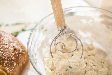 Wooden bread whisk in a bowl of bread dough with fresh bread loaf beside it