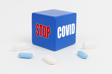 On a white surface are pills and a blue cube that says - STOP COVID