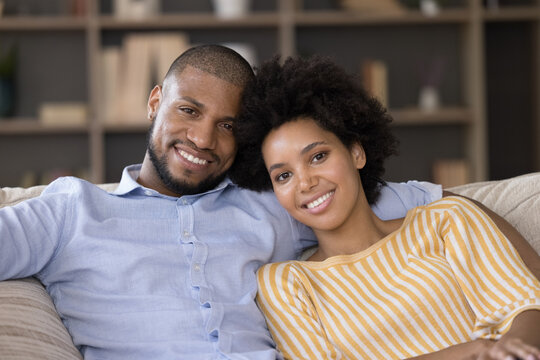 Beautiful young 35s African good-looking couple in love relaxing sitting on cozy sofa smiling looking at camera. Close up of happy homeowners family portrait. Romantic relationships, dating concept