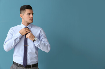 Young handsome man in formal clothes looking aside and adjusting tie on color background