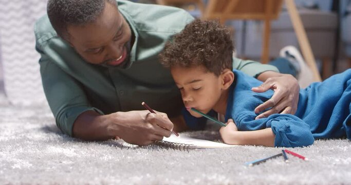 Close up of happy African American man hugging his beloved grandson and spending time lying on the floor drawing picture for him.