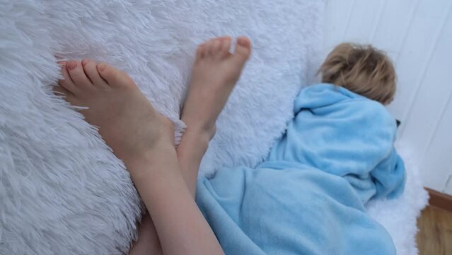 The teenage girl sat comfortably on the couch. The child is dressed in a blue house robe. In the foreground are bare children's feet and a fluffy blanket. The child plays in the smartphone. Social