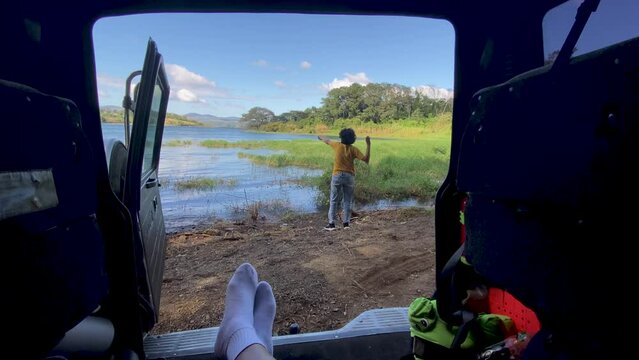 hombre acostado dentro de su veh&iacute;culo, relajado viendo a su hijo lanzar piedras a un lago 
