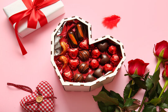Heart shaped box with tasty chocolate candies, roses and gift on pink background