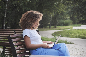 Modern African American woman focused remotely working use laptop sitting on bench at summer park. Female freelancer enjoying distance online communication chatting surfing internet on computer