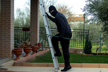 thief dressed in black with a crowbar climbing a ladder trying to sneak into a house © gianni