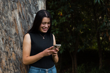 Una joven sonriente con el pelo largo y negro parada cerca de una pared de roca mirando  su móvil en las manos - obrazy, fototapety, plakaty
