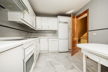 Kitchen with walls covered with white wooden cabinets, white countertops and gray tiles, white marble floor and white wooden table and pine door