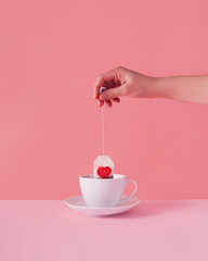 Immersing paper compound  tea bag with red heart into white cup on pastel pink background. ST...