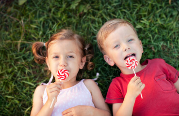 a smiling boy and a girl are lying on the grass and eating a lollipop on a stick
