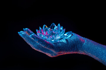 Woman holding fragile crystal lotus flower. Hand covered with shining glitter under neon colored...