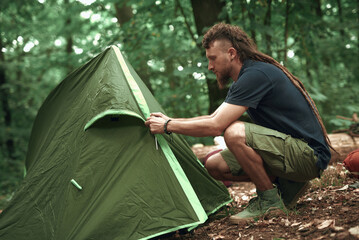 Young hippie hiker setting up his tent in the forest, pitching in a forest clearing, getting ready for a camping sleepover - obrazy, fototapety, plakaty