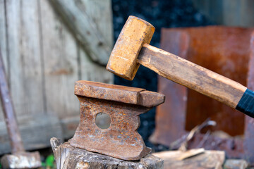 Old rusty anvil stands on a stump in the backyard of village house, sledgehammer leaning on wooden fence enclosing a barn with coal on blurred background. Heavy working tools