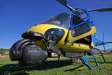 An unmarked news helicopter, outfitted with a powerful camera at the nose, is shown in a field during a sunny day.