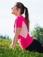 Smiling adult woman in pink T-shirt is practicing set of stretching exercises on nature.