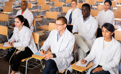 Multiethnic group of people in white lab coats listening carefully and making notes during professional training for health workers