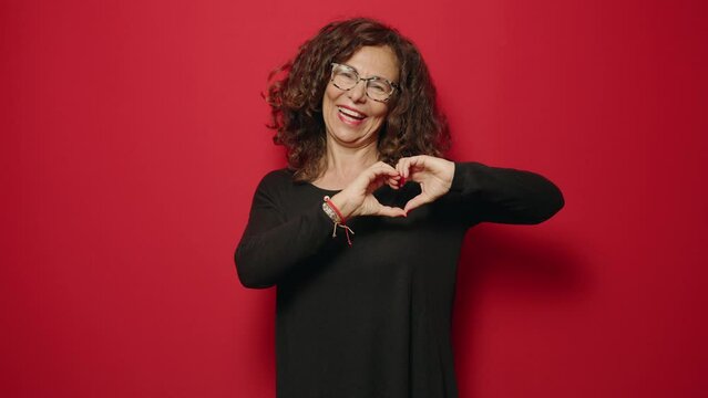 Middle age woman smiling confident doing heart symbol with hands over red background