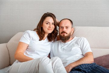 Obraz na płótnie Canvas Young beautiful couple in pajamas hug while sitting on couch at cozy home interior. Married man and woman romantic relationship. Husband and wife portrait, morning routine