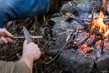 A man's hand's whittling a stick with a survival knife by a campfire.
