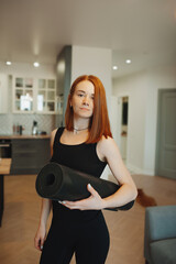 portrait young woman with bright red hair in sportswear with training mat in interior of stylish house. home workouts. online sports. vertically, selective focus