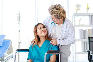 Obraz premium Portrait shot of senior professional female doctor in white lab coat with stethoscope standing smiling help pushing disabled handicapped injury woman patient in hospital uniform sitting on wheelchair