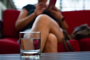 A selective focus shot of a young lady is preoccupied with her phone while relaxing on a comfy couch with a glass of refreshing water on the table in front of her - obrazy, fototapety, plakaty