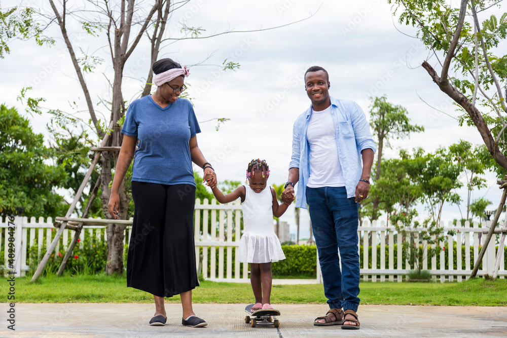 Wall mural Happy African American parent teach his little daughter to play skateboard in the public park during summer for family leisure time concept.