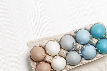 Easter eggs pastel colored in egg box decorated flowers on white wood background. Minimal Easter  concept, dyed chicken egg with color gradient from blue to beige coloured in carton pack
