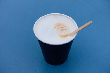 A cup of hot coffee with milk in a paper cup stands on a blue table. White foam on coffee with a...