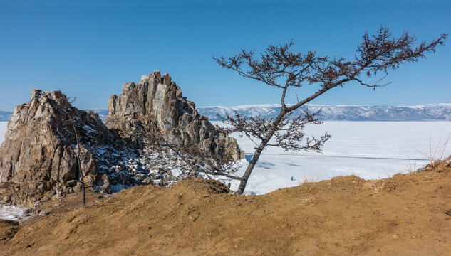 A picturesque double-headed rock against the blue sky. Steep cracked granite slopes. A tiny silhouette of a man is visible on a frozen lake. Bare trees in the foreground. Baikal. Shamanka Mountain